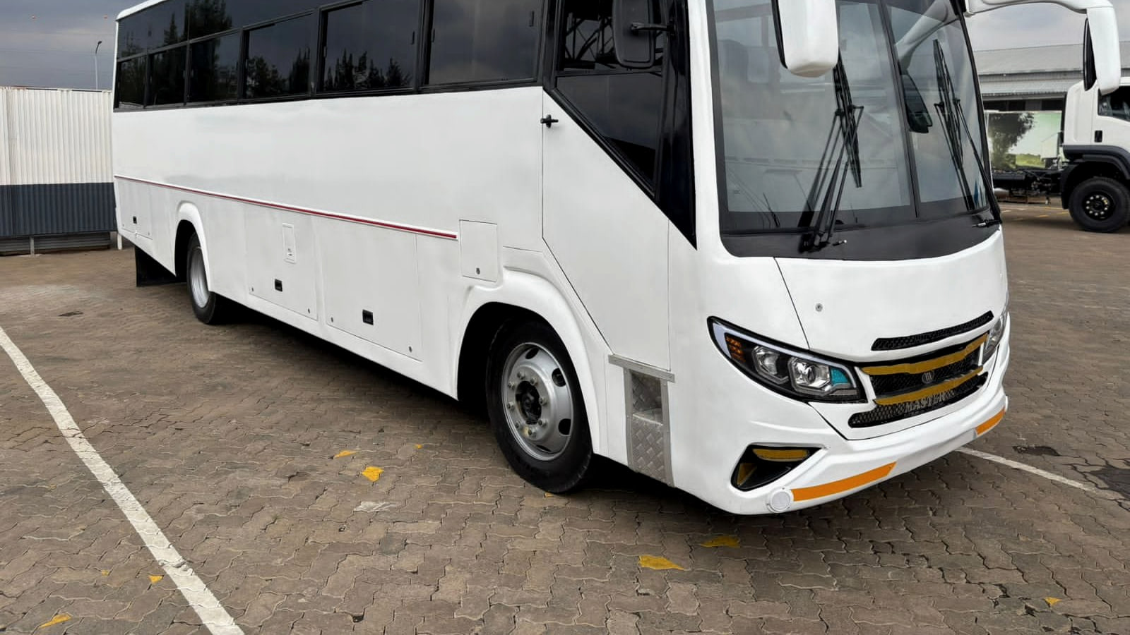 Isuzu NQR81M Zuri, a 29-seat coach in white with a red waistline stripe, front three-quarter view on a paved yard.